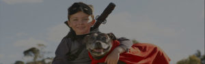 Child and dog in superhero costumes outdoors, blue sky background.